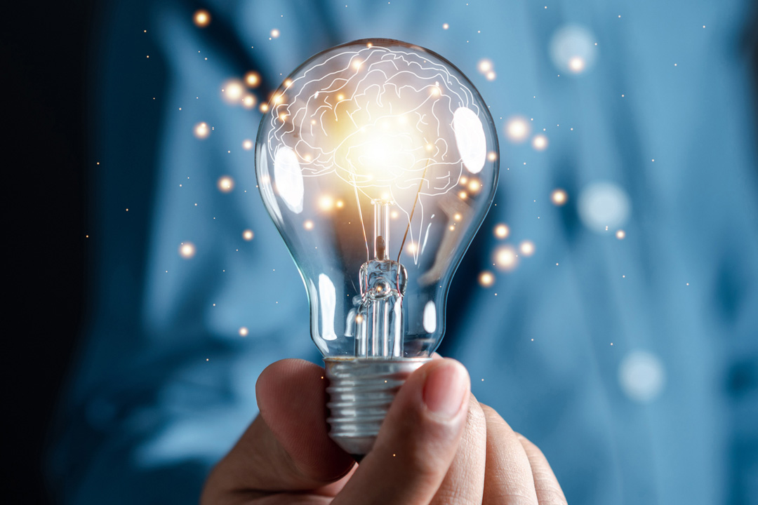 Hand holding a glowing lightbulb containing a brain illustration, surrounded by sparkling particles