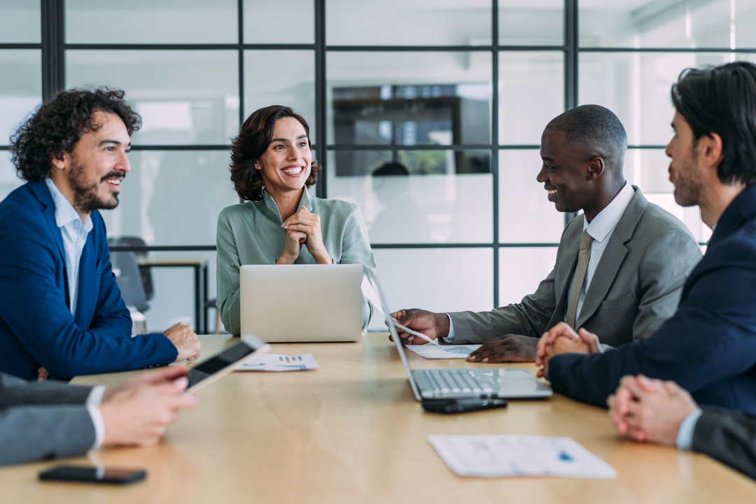 Four colleagues smiling around a conference table with laptops and documents in a bright glass-walled office