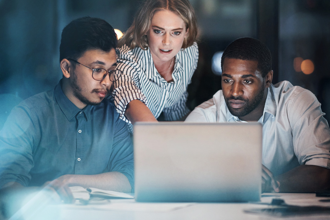 Three colleagues intently studying a laptop screen together in a dimly lit workspace at night